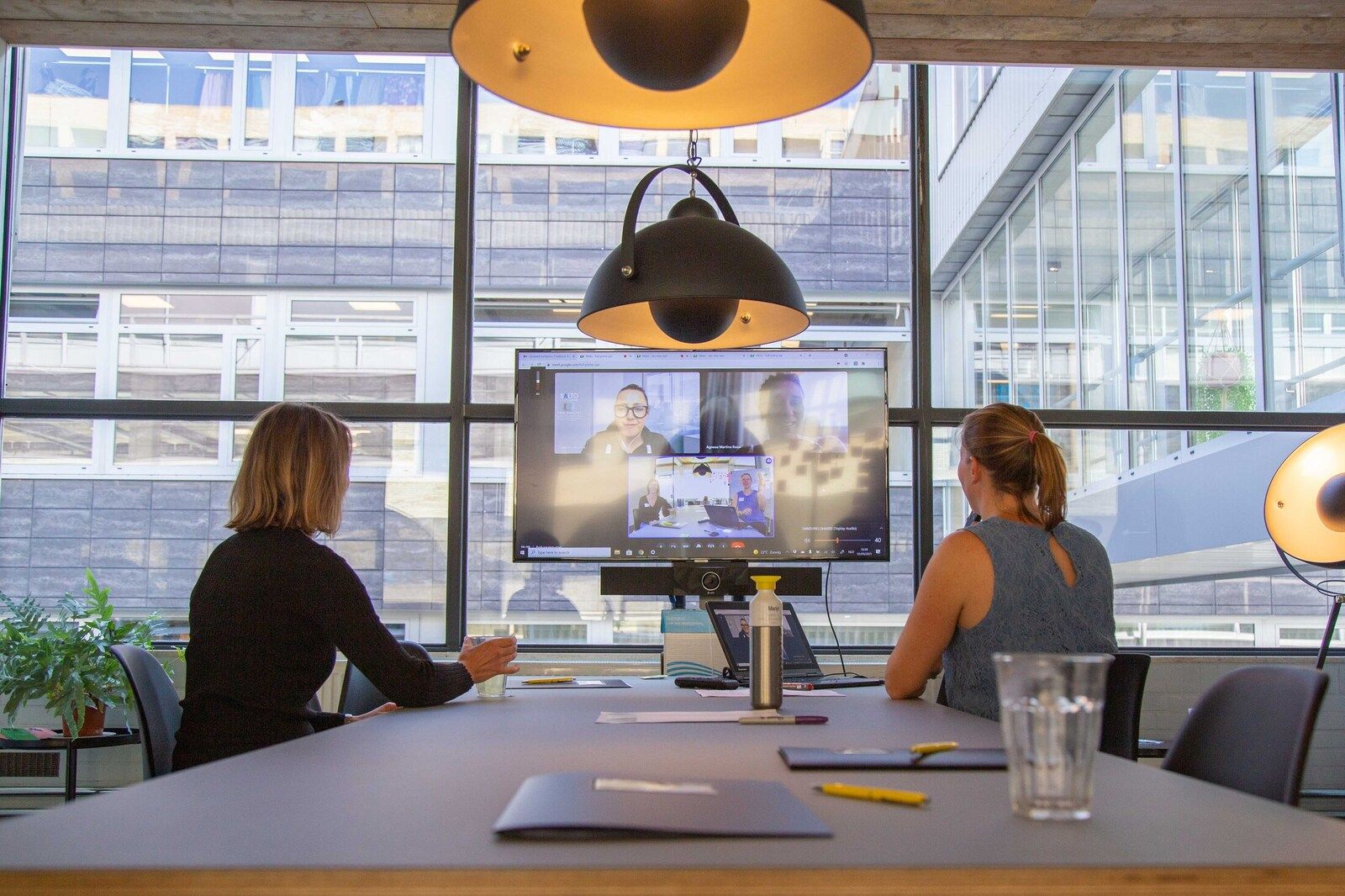 2 personen aan vergadertafel overleggen met 3 personen op een groot scherm